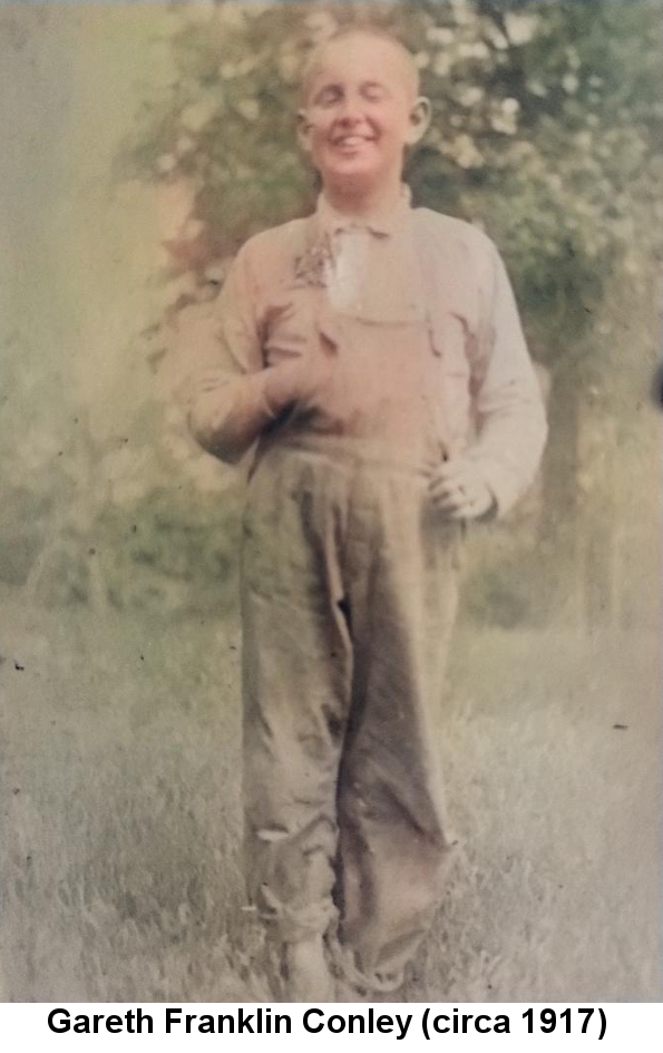 Colorized black and white photo of Gareth Franklin Conley, about ten years old, with close-cropped blond hair, smiling with eyes nearly closed, his right hand inserted between the buttons of his shirt, standing in a grassy yard in front of a tree